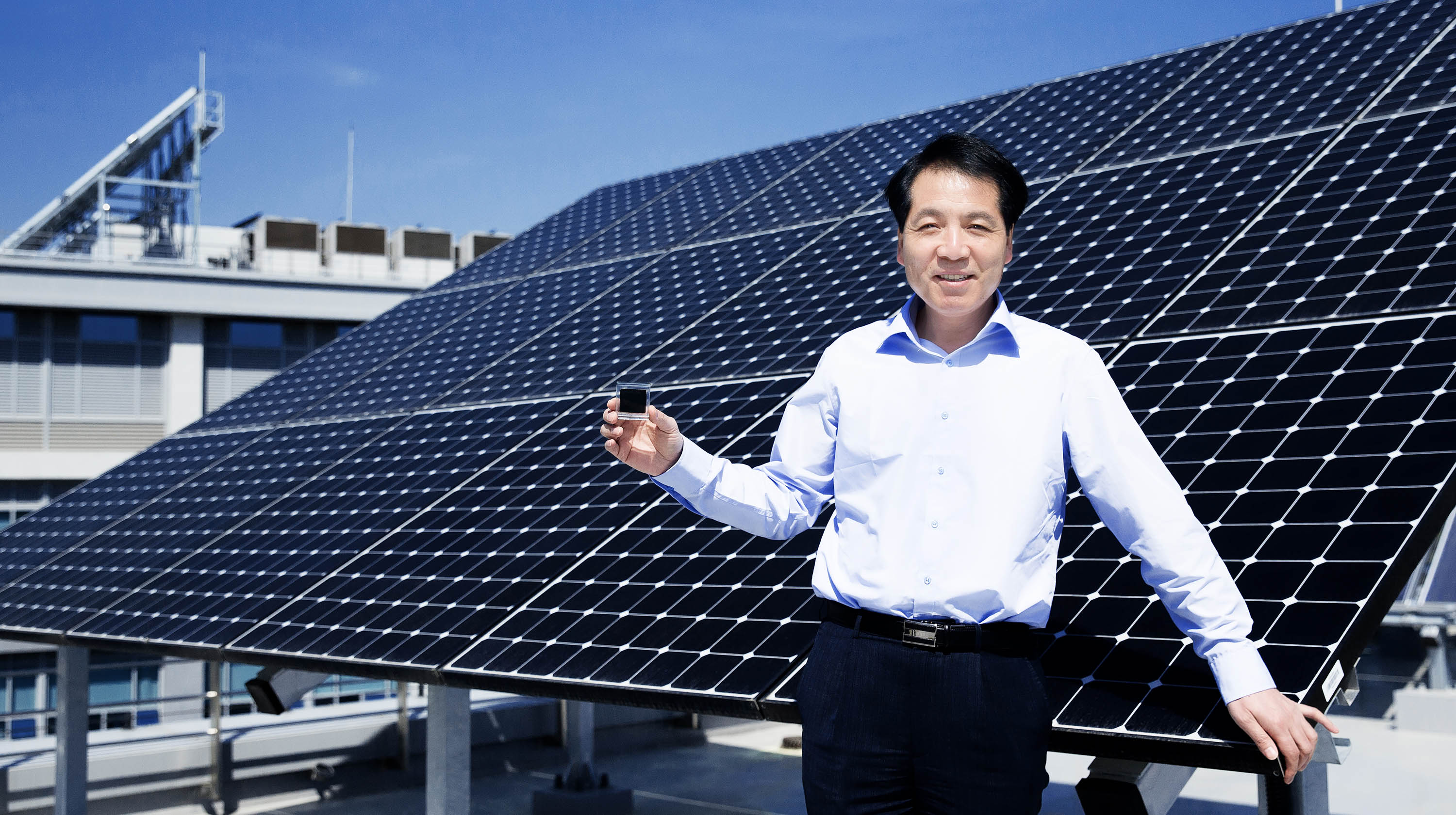 석상일 교수가 UNIST 공학관 옥상에 설치된 실리콘 태양전지 판을 배경으로 서 있다. 그의 손에는 차세대 태양전지의 강력한 후보인 페로브스카이트 태양전지 셀이 들려있다. | 사진: 김경채